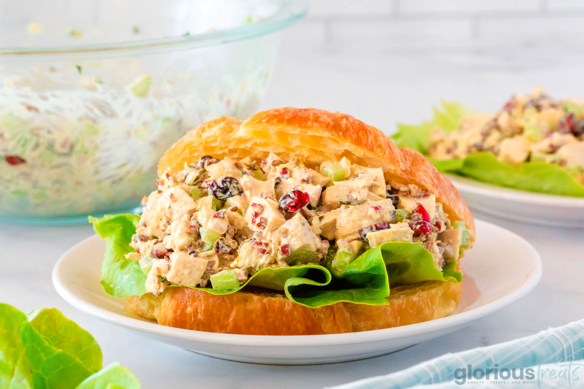 Chicken salad sandwich made with croissant and lettuce on small white round plate. Bowl of chicken salad can be seen in the background.