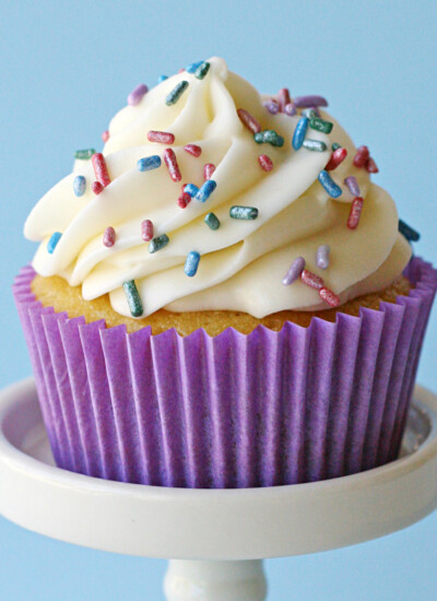 single vanilla cupcake on white cupcake stand with purple liner and pastel sprinkles