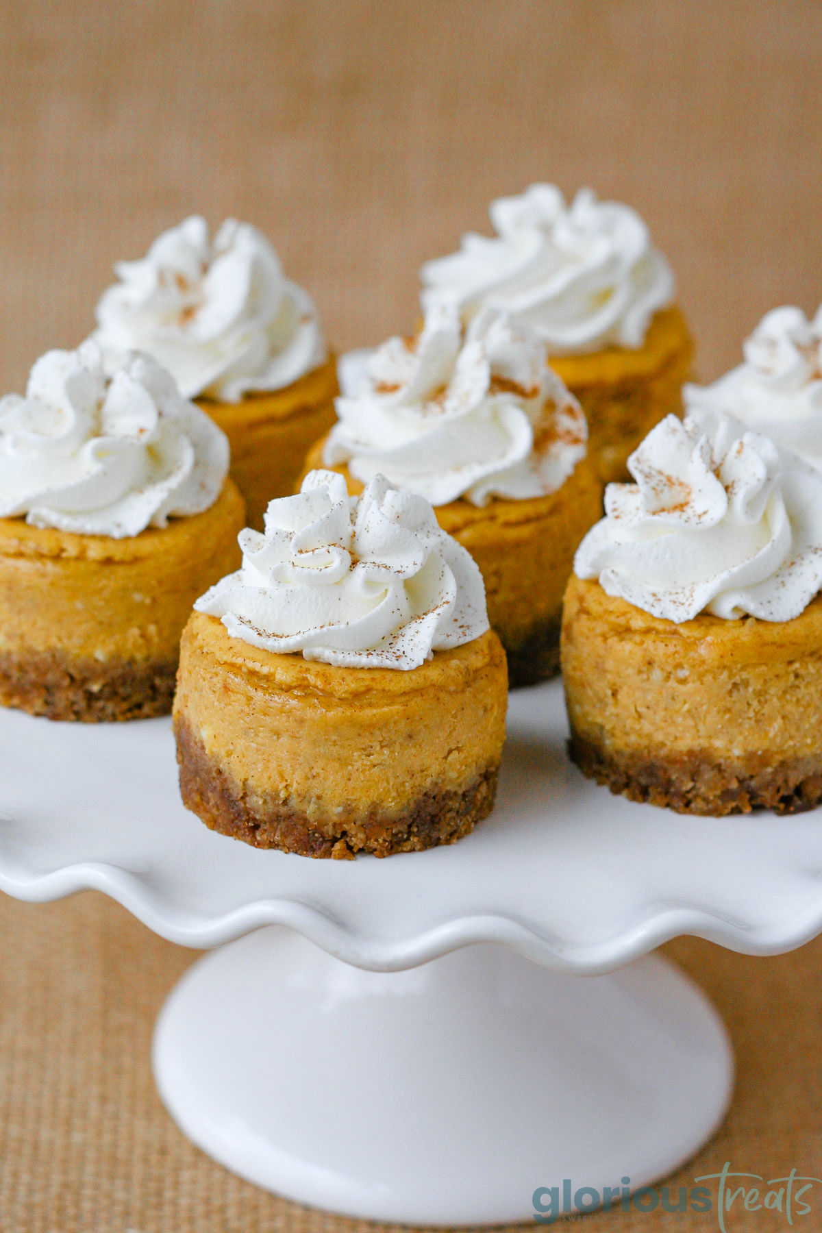 White scalloped cake stand topped with mini pumpkin cheesecakes topped with whipped cream. Burlap sack is the backdrop.