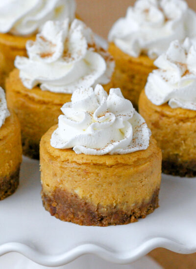 Close up shot of white scalloped cake stand topped with mini pumpkin cheesecakes topped with whipped cream. Burlap sack is the backdrop.