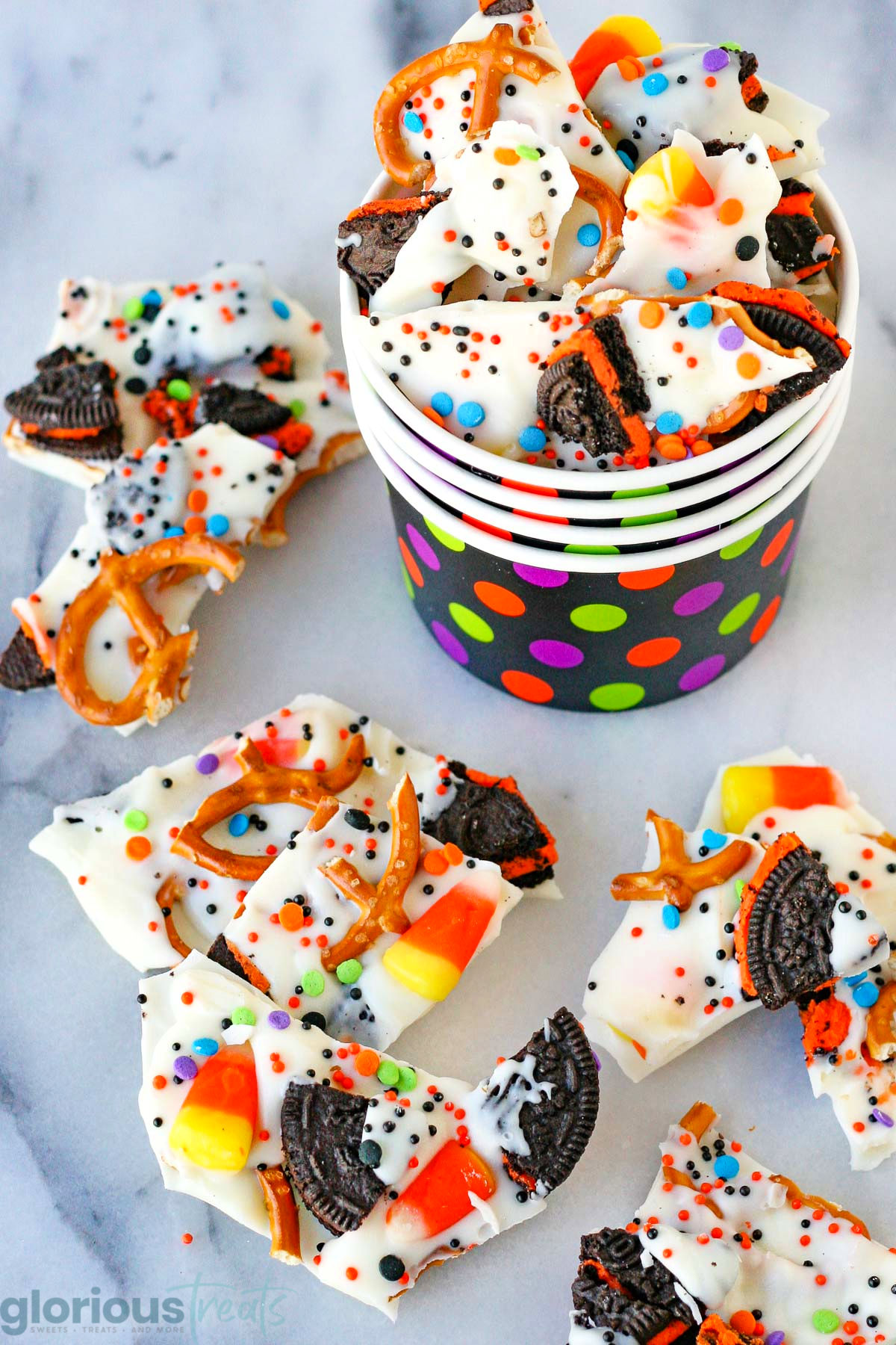 Halloween Bark broken into bite sized pieces and placed in colorful halloween cup filled to the top. Made with pretzels, sprinkles and halloween cookies and candy. More bark can be seen scatter about the base of the cup.