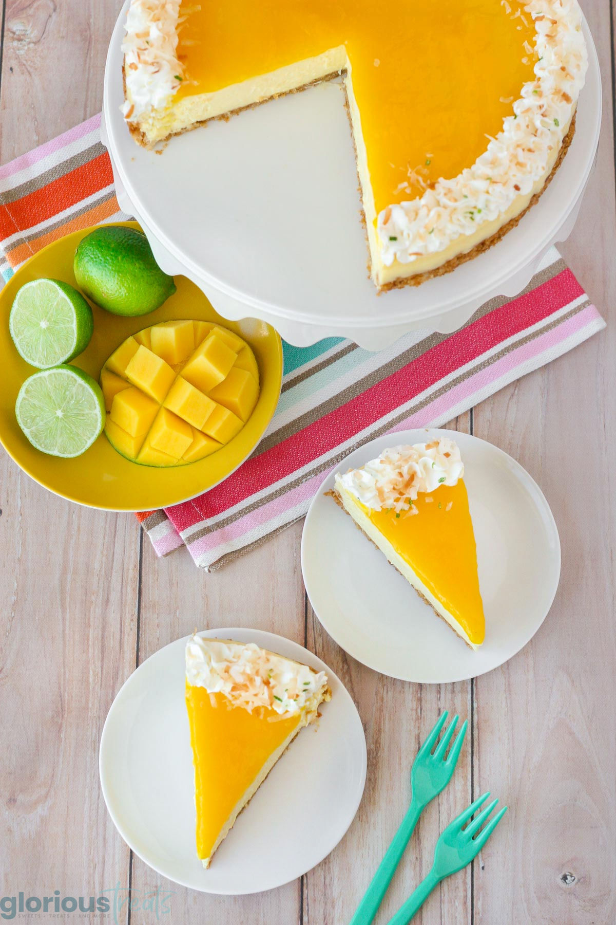 Two slices of lime mango cheesecake on white round plates and the rest of the cheesecake is on a white cake stand.