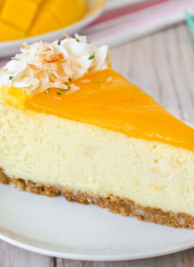 Slice of mango lime cheesecake on a round white plate. Another slices and fresh mango and limes can be seen in the background.