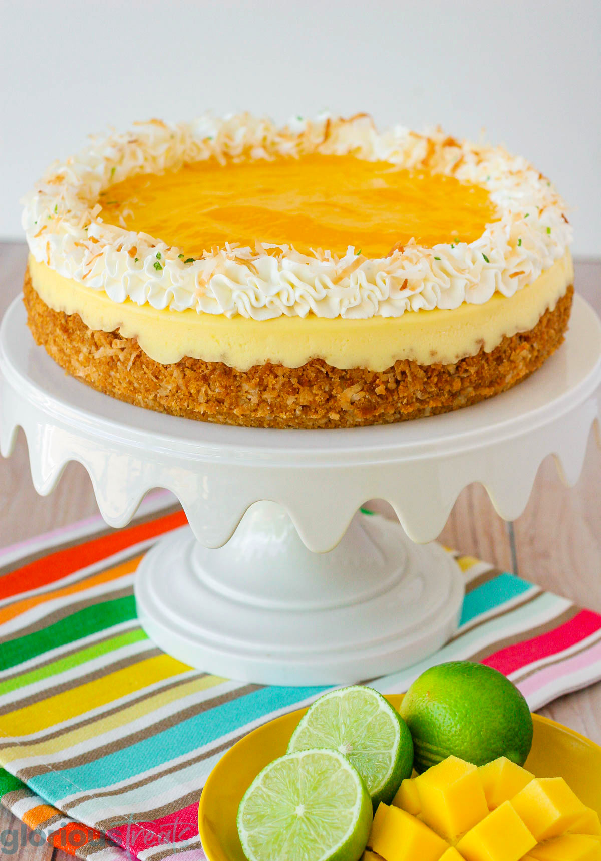 Whole mango lime cheesecake on a white cake stand sitting on a colorful towel next to sliced mango and lime. 