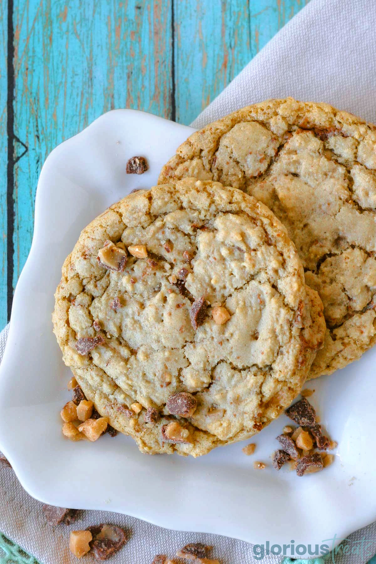 Two toffee cookies on a white scalloped square plate with toffee pieces scattered nearby.