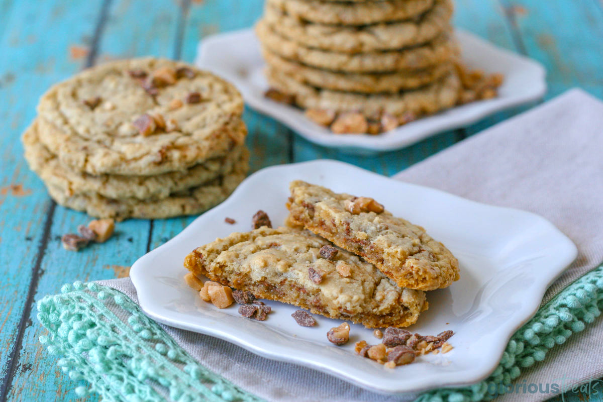 Toffee cookie broken in half on white plate. Stack of more toffee cookies can be seen in the background.