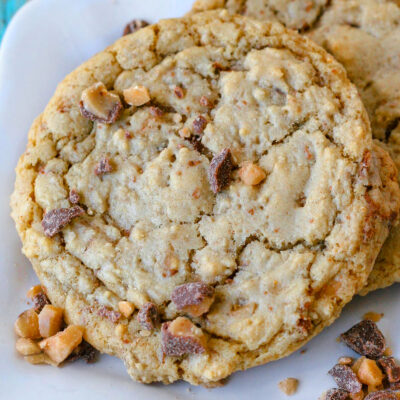 Two toffee cookies on a white scalloped square plate with toffee pieces scattered nearby.