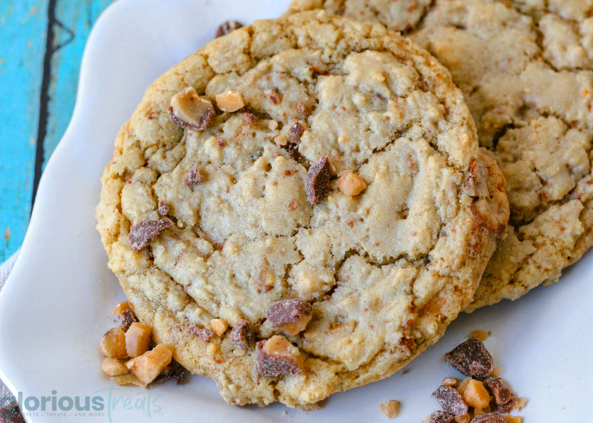 Two toffee cookies on a white scalloped square plate with toffee pieces scattered nearby.