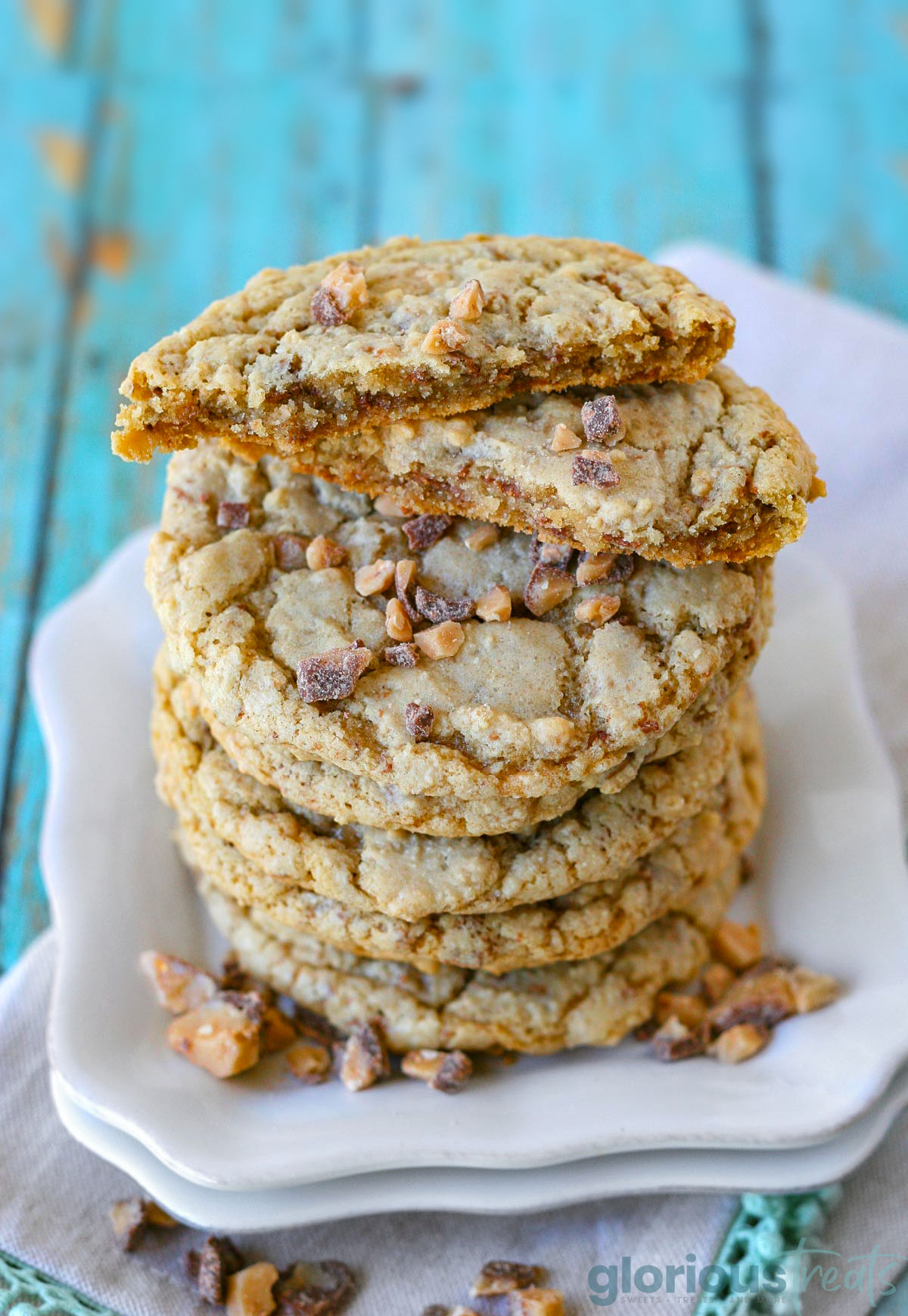 Toffee cookies stacked high on a square white plate. Top cookie has been broken in half to show the chewy interior.