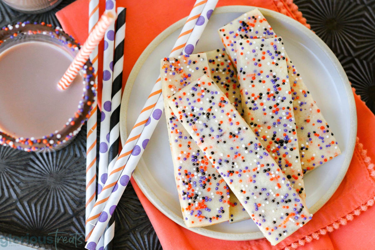 Top down shot of plate of halloween cookies with sprinkles, colorful striped straws and a glass of chocolate milk.