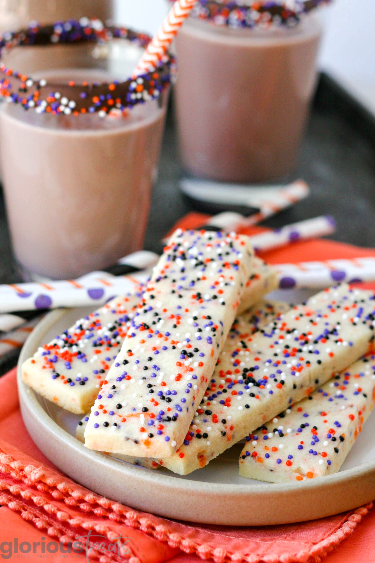 Halloween sprinkle cookies on white round plate with glasses of chocolate milk in the background. Striped paper straws are behind the cookies and the plate sits on an orange cloth napkin.