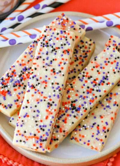 Halloween sprinkle cookies on white round plate with glasses of chocolate milk in the background. Striped paper straws are behind the cookies and the plate sits on an orange cloth napkin.