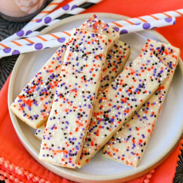 Halloween sprinkle cookies on white round plate with glasses of chocolate milk in the background. Striped paper straws are behind the cookies and the plate sits on an orange cloth napkin.