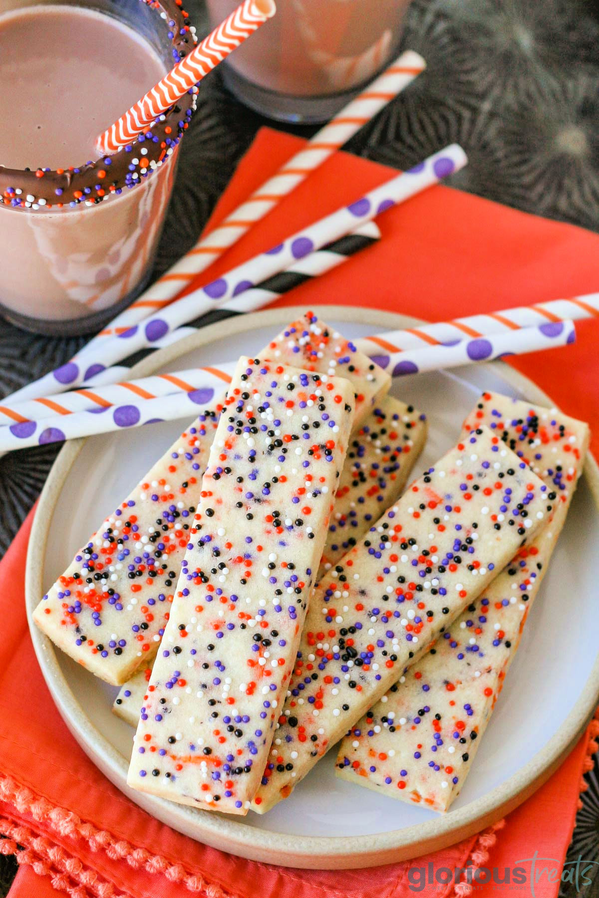 Halloween sprinkle cookies on white round plate with glasses of chocolate milk in the background. Striped paper straws are behind the cookies and the plate sits on an orange cloth napkin.