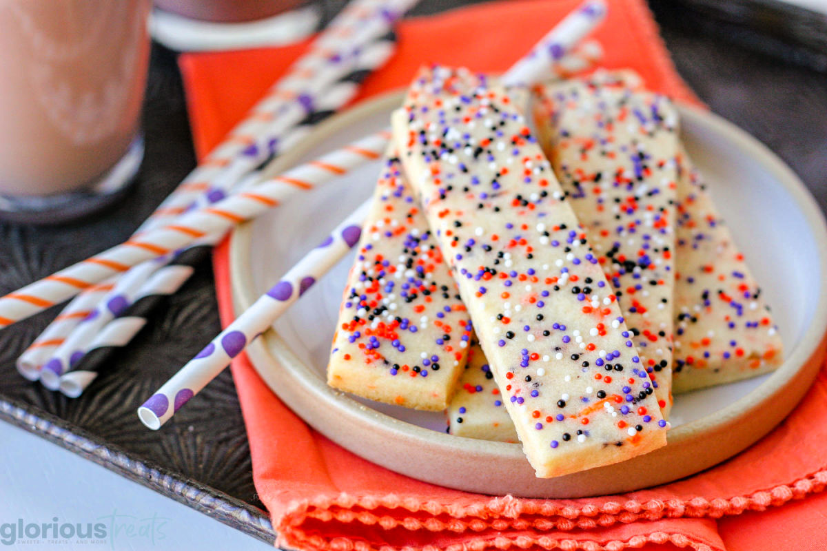 Wide shot of halloween sprinkle sugar cookies on round plate sitting on orange cloth napkin next to colorful striped straws.