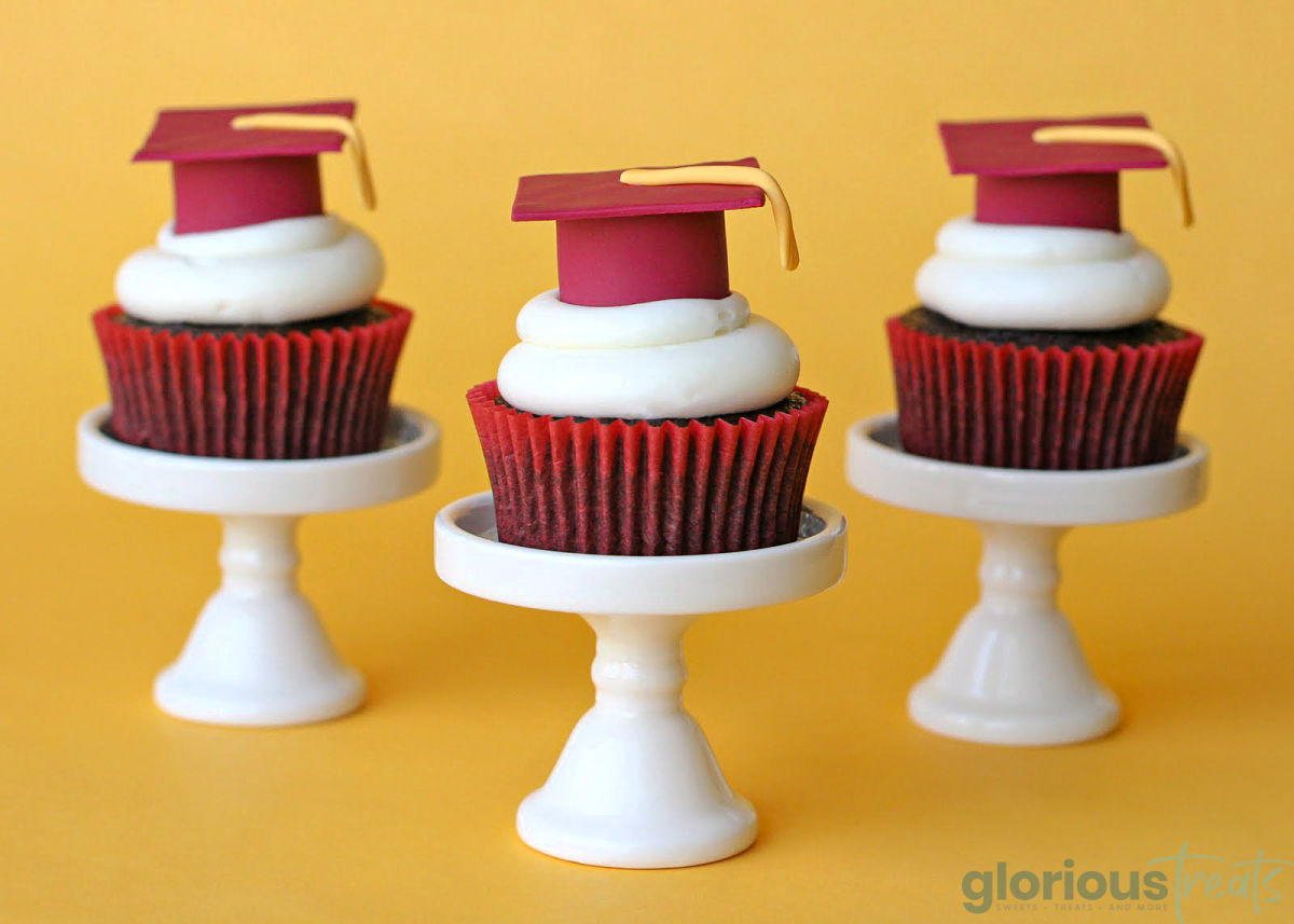 three graduation cupcakes on individual white cupcake stands with red fondant graduation caps on them. 