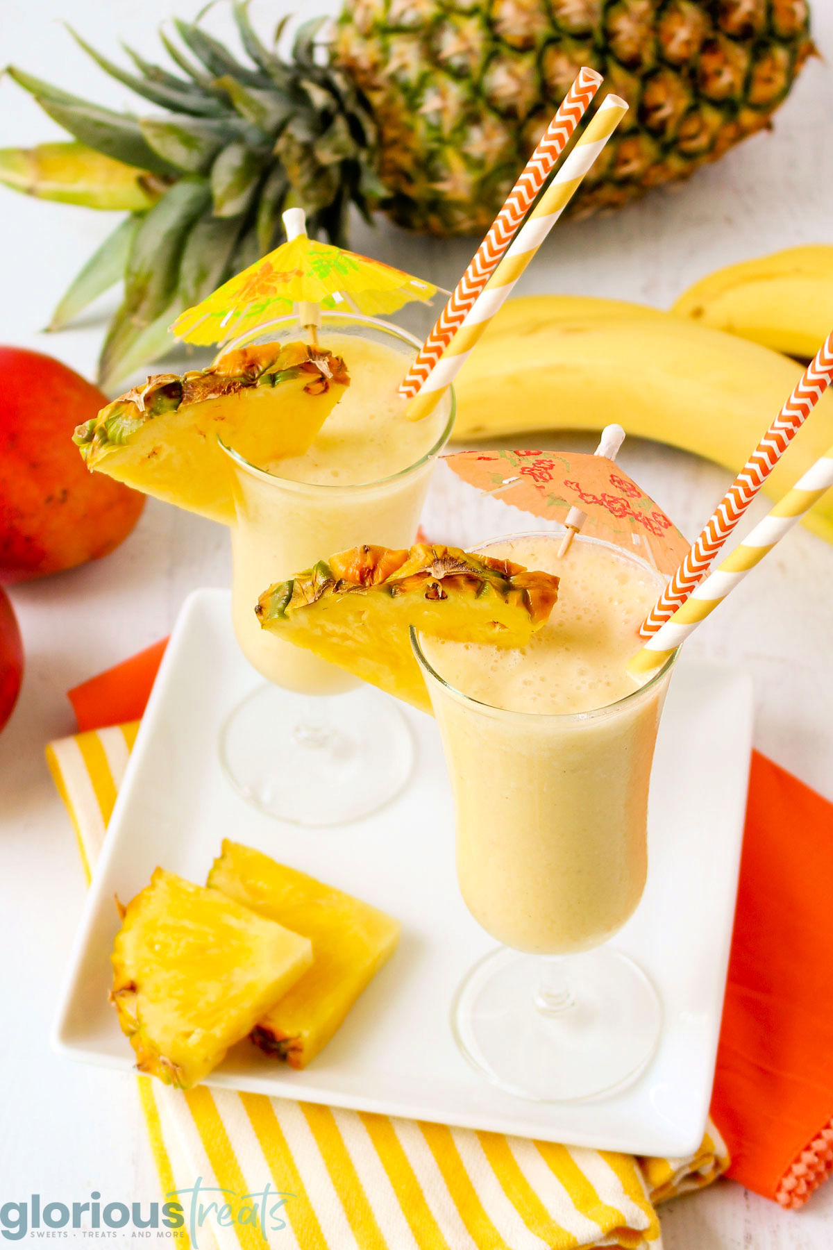 angled look down at two tall glasses filled with a tropical smoothie recipe sitting on a white tray garnished with wedges of pineapple. A whole pineapple and bananas can be seen in the background.