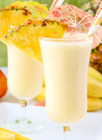 two light yellow fruit smoothies garnished with pineapple slices, paper umbrellas, and paper straws.