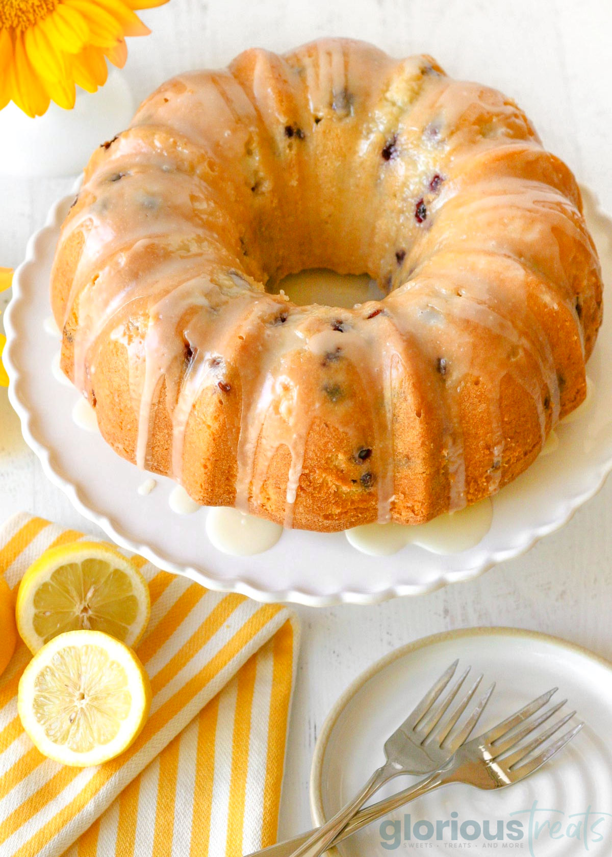 overhead shot of a lemon blueberry bundt cake drizzled in icing on a white serving plate with lemons in the bottom left corner