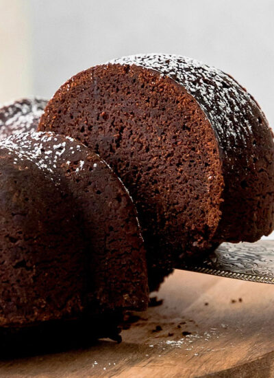 a chocolate bundt cake on a wood cutting board with a spatula removing a slice of cake from it.