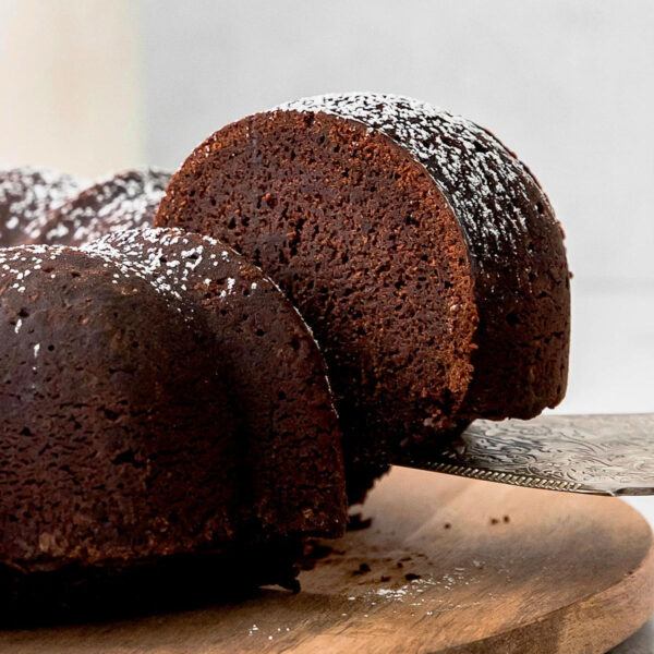 a chocolate bundt cake on a wood cutting board with a spatula removing a slice of cake from it.