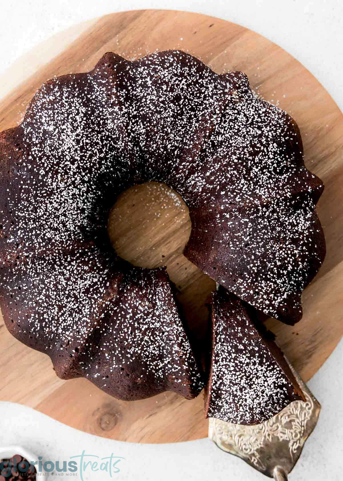 an overhead shot of chocolate bundt cake on a wooden cutting board. a spatula is removing a slice of cake.
