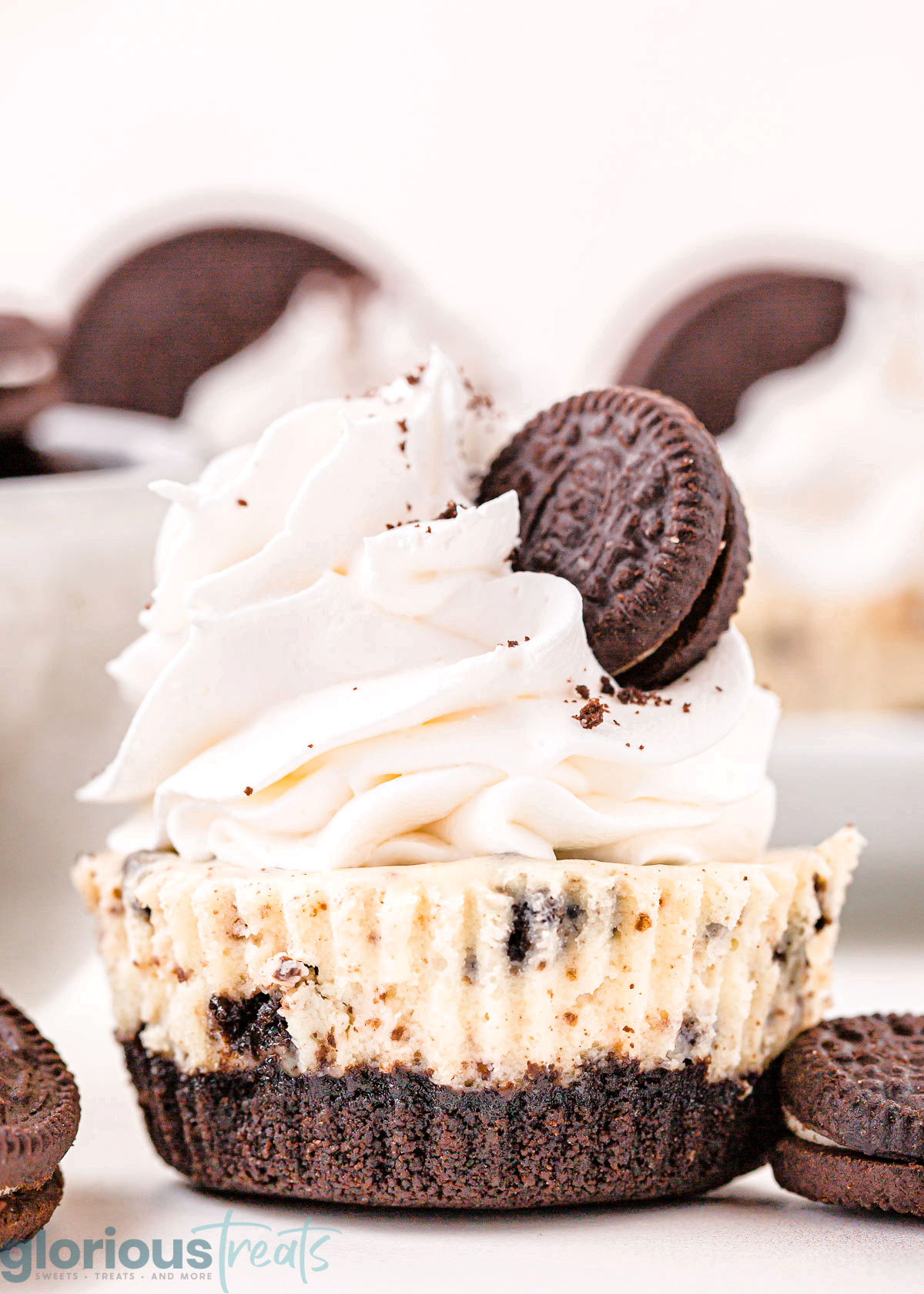 mini oreo cheesecake topped with whipped cream and a mini oreo sitting on a white surface with oreo cookies scattered about.