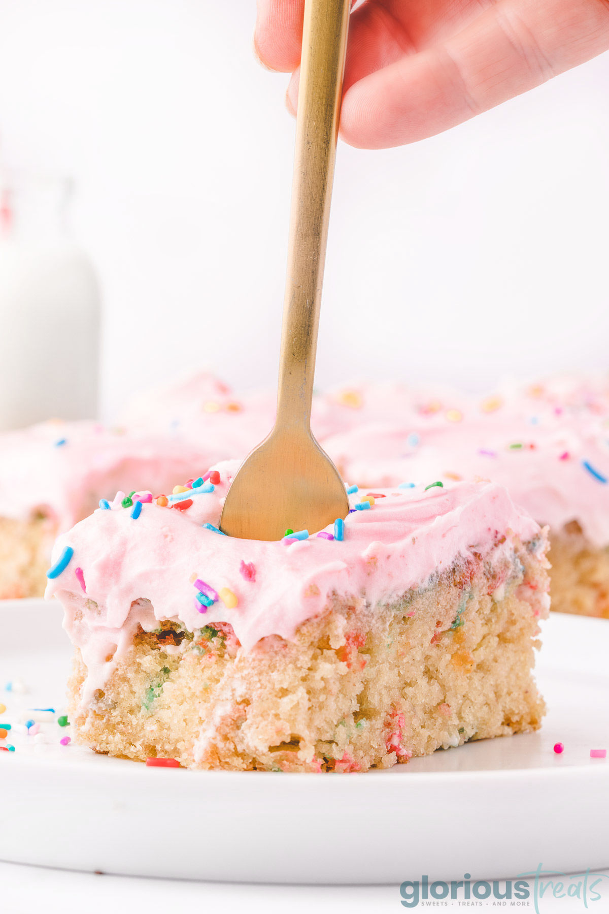a hand holding a gold fork in a slice of funfetti sheet cake on a white plate.