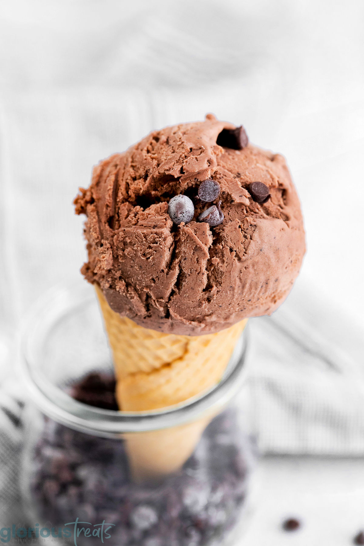 A scoop of no-churn chocolate ice cream on a cone sitting in a glass jar full of chocolate chips.