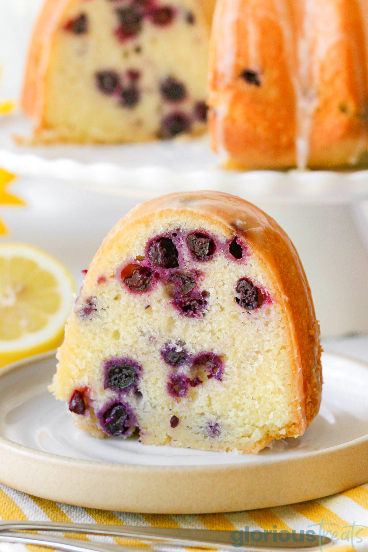 a slice of blueberry lemon bundt cake on a white plate with the full cake in the background