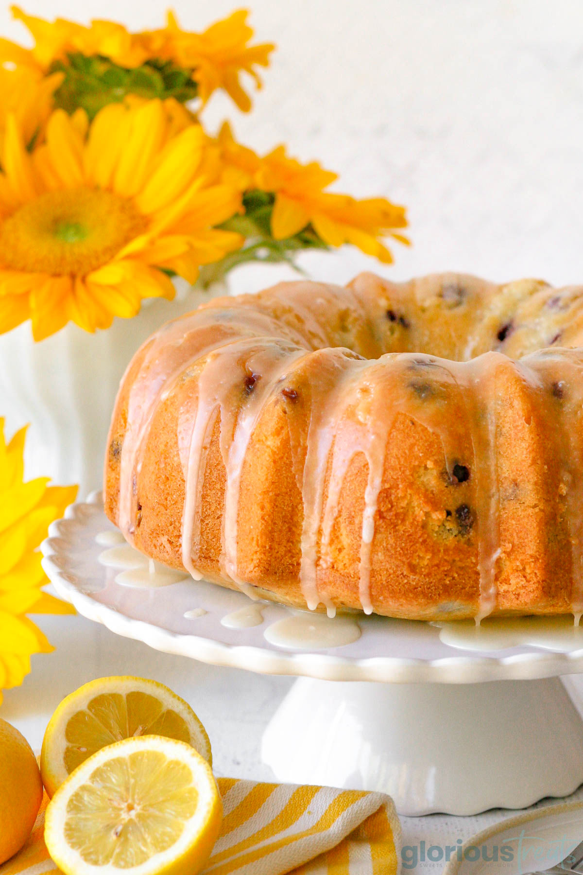 a full lemon blueberry bundt cake drizzled with glaze and served on a white cake plate. there are yellow flowers in the left back corner