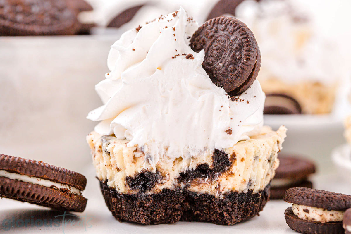 mini Oreo cheesecake with a bite taken out of it so you can see the cookies and creamy filling. The cheesecake has been topped with whipped cream and a tiny Oreo cookie.