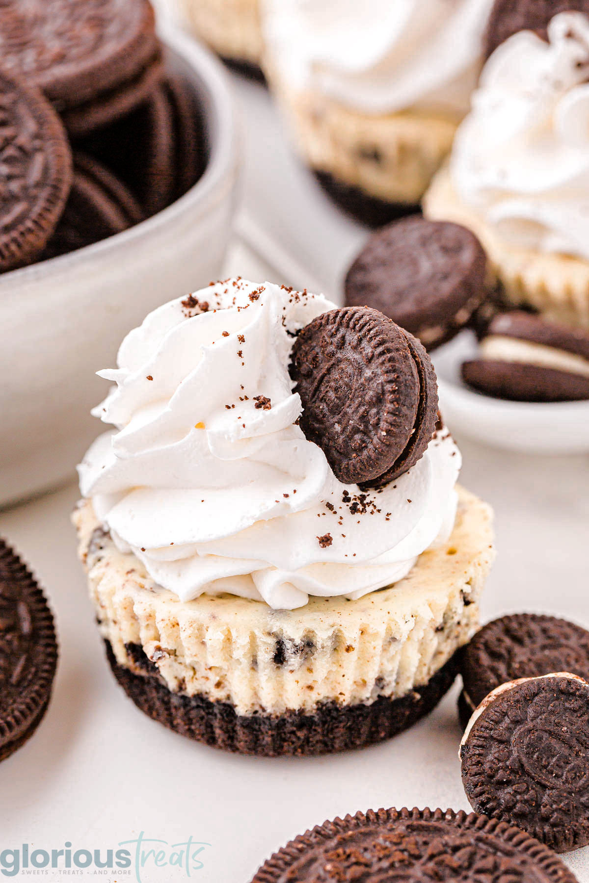 mini oreo cheesecake topped with whipped cream and a mini oreo sitting on a white surface with oreo cookies scattered about.