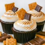 three chocolate s'mores cupcakes topped with a hershey square and graham crackers and extra toppings on the white surface beneath them.