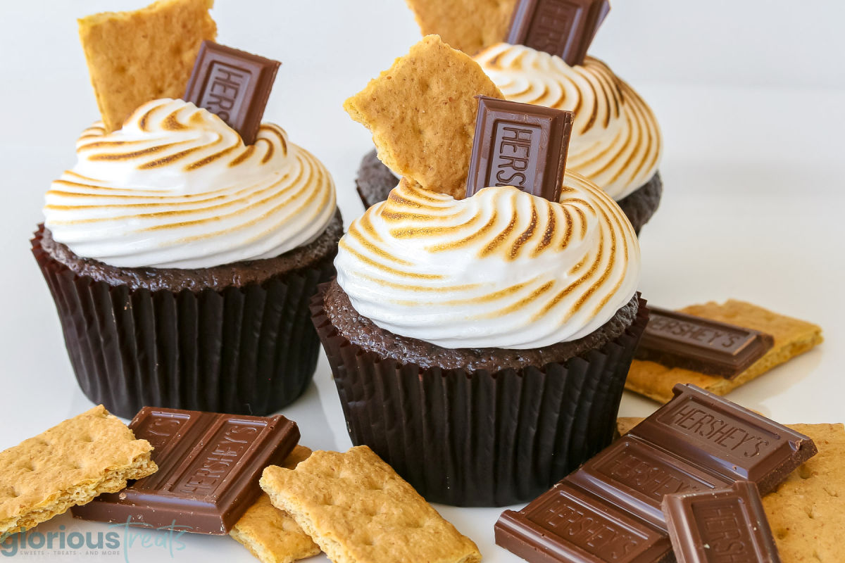 three chocolate s'mores cupcakes topped with a hershey square and graham crackers and extra toppings on the white surface beneath them.