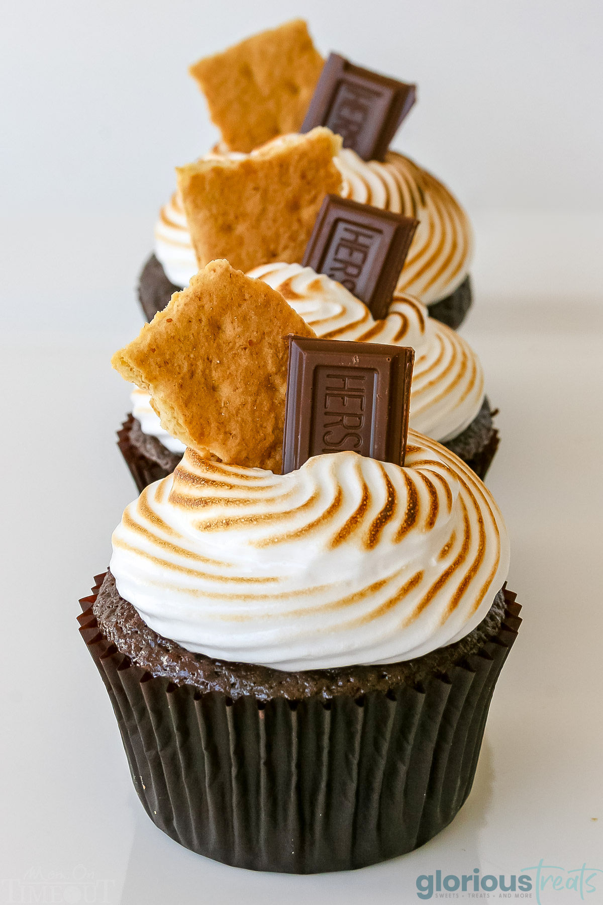 Three smores cupcakes lined up in a row on a white surface. Cupcakes have been topped with marshmallow frosting, graham crackers and chocolate candy.