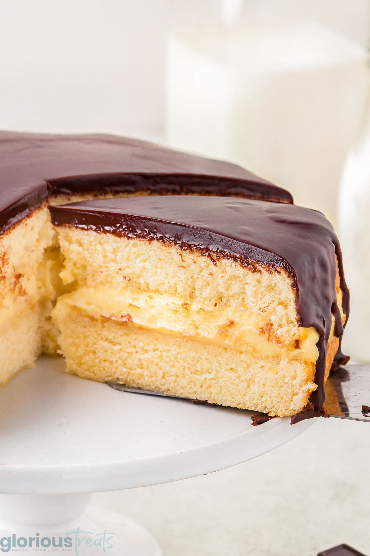 Boston Cream Pie on a white cake stand with multiple slices removed and one more ready to be served. 