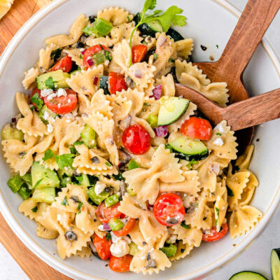 Greek Pasta Salad in a white bowl with wood serving spoons sticking out of it.