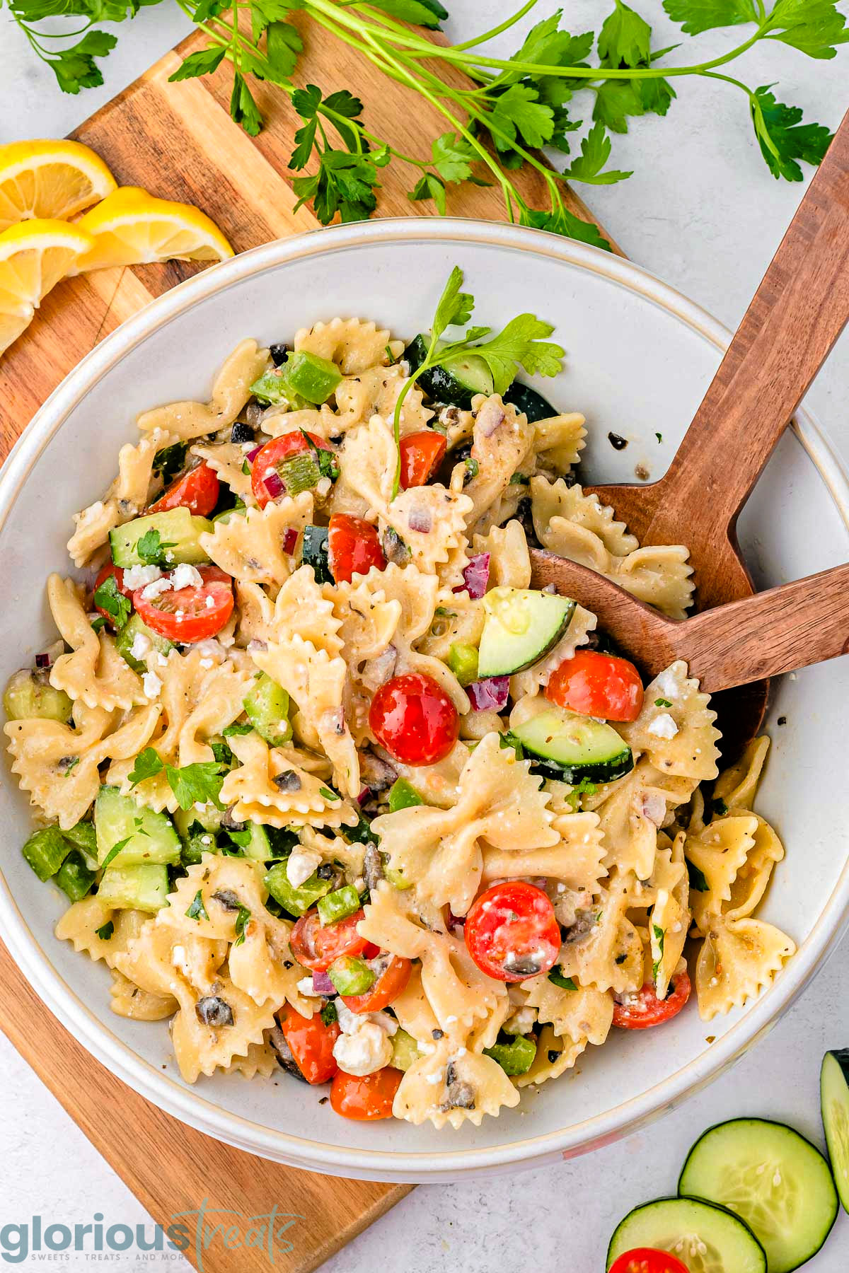 Greek Pasta Salad in a white bowl with wood serving spoons sticking out of it.