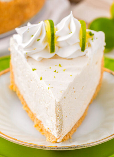 Slice of key lime cheesecake on a white round plate sitting on a larger green plate. Cheesecake slice is topped with whipped cream and lime slices.