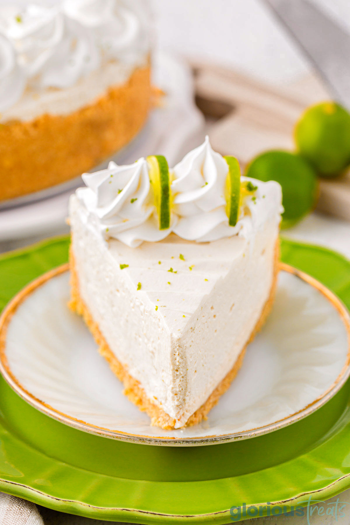 Slice of key lime cheesecake on a white round plate sitting on a larger green plate. Cheesecake slice is topped with whipped cream and lime slices.