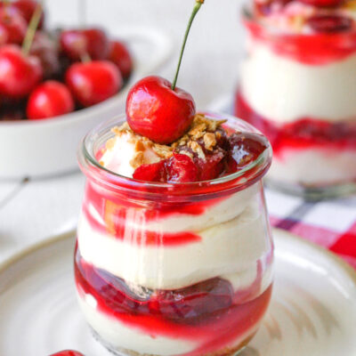 a jar of no bake cherry cheesecake on a white plate with another jar and a bowl full of cherries out of focus in the background