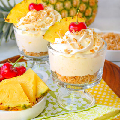 two small glasses of pina colada mousse beside a dish of fresh pineapple and cherries, with a whole pineapple on its side in the background