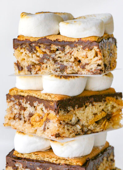 Three decadent smores rice krispie treats stacked on a log. Looking straight on so the layers of rice krispie treats, chocolate, graham crackers, and toasted marshmallows are visible.