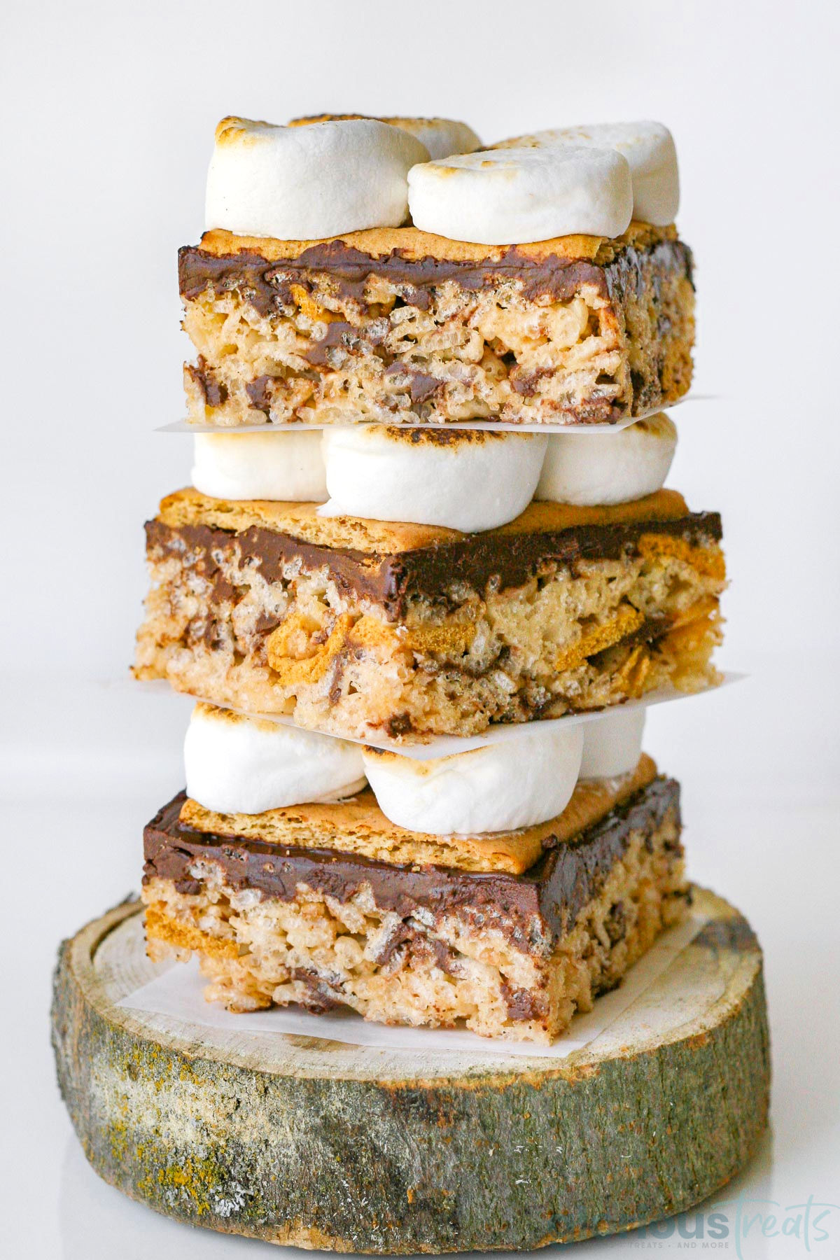 Three decadent smores rice krispie treats stacked on a log. Looking straight on so the layers of rice krispie treats, chocolate, graham crackers, and toasted marshmallows are visible.