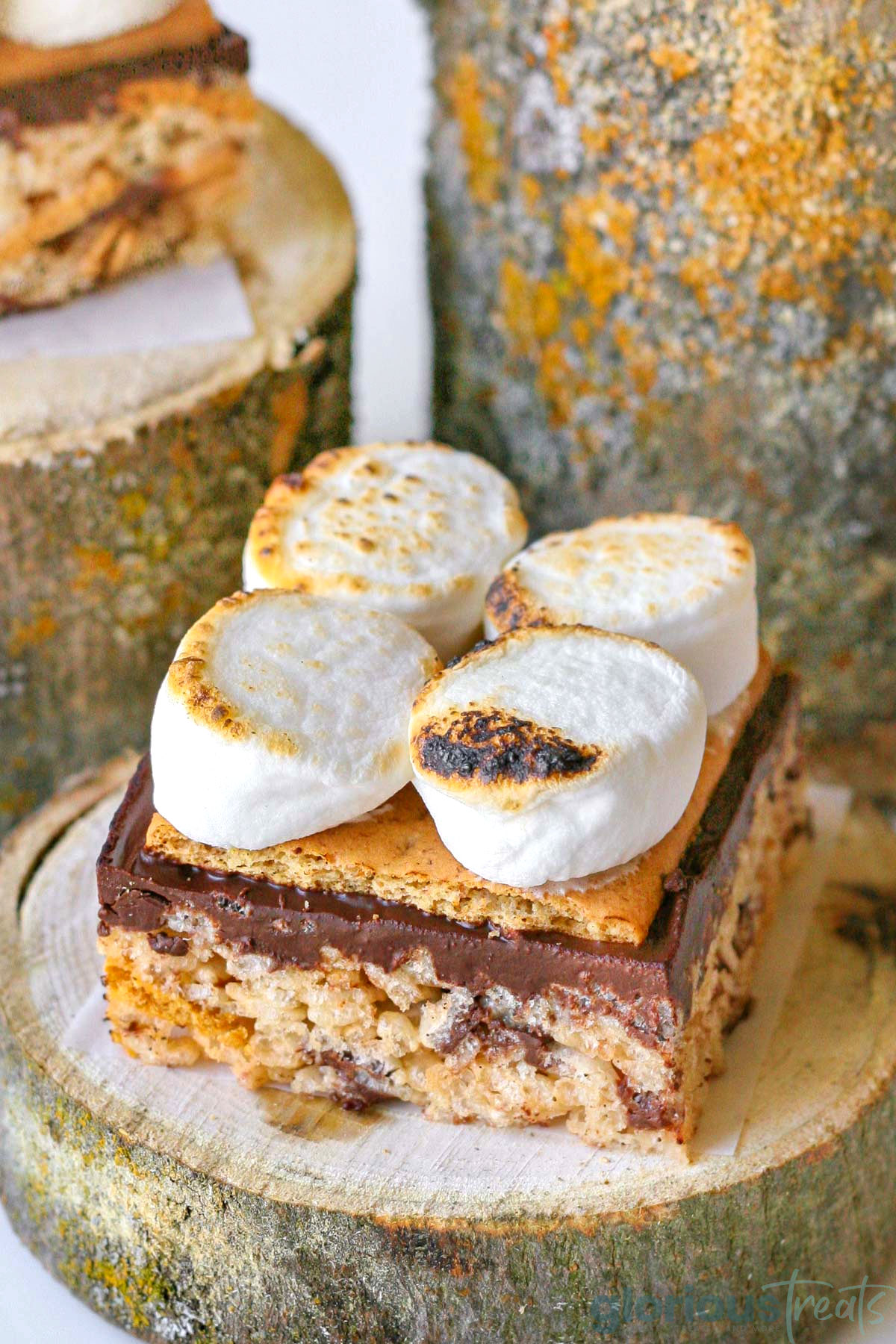 Toasted marshmallows on a smores rice rice krispie treat sitting on a log. More logs of varying heights can be seen in the background.
