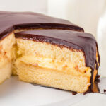 A slice of boston cream pie held on a spatula over a white cake stand.