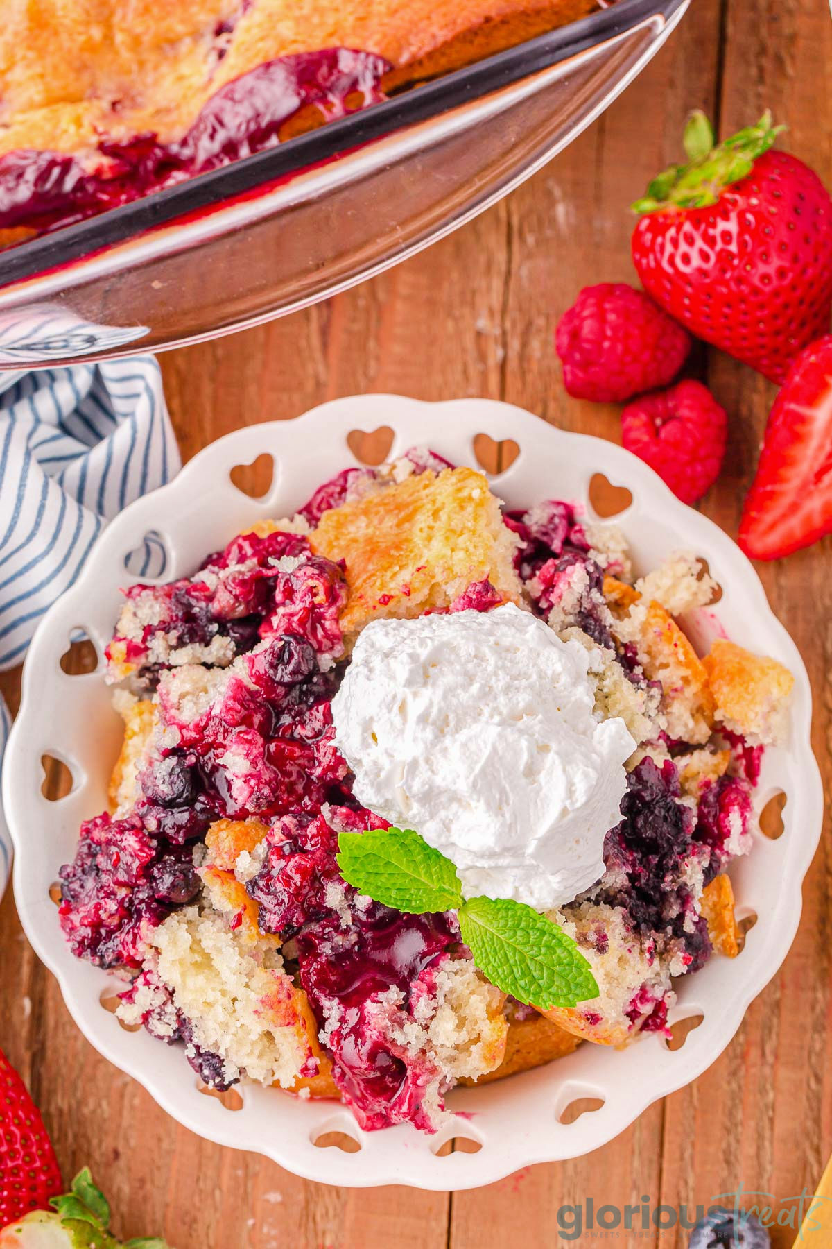 Top down view of berry cobbler recipe in a white bowl with heart cutouts along the edge and is topped with whipped cream and a mint sprig. The bowl is sitting next to the baking dish with the rest of the cobbler.