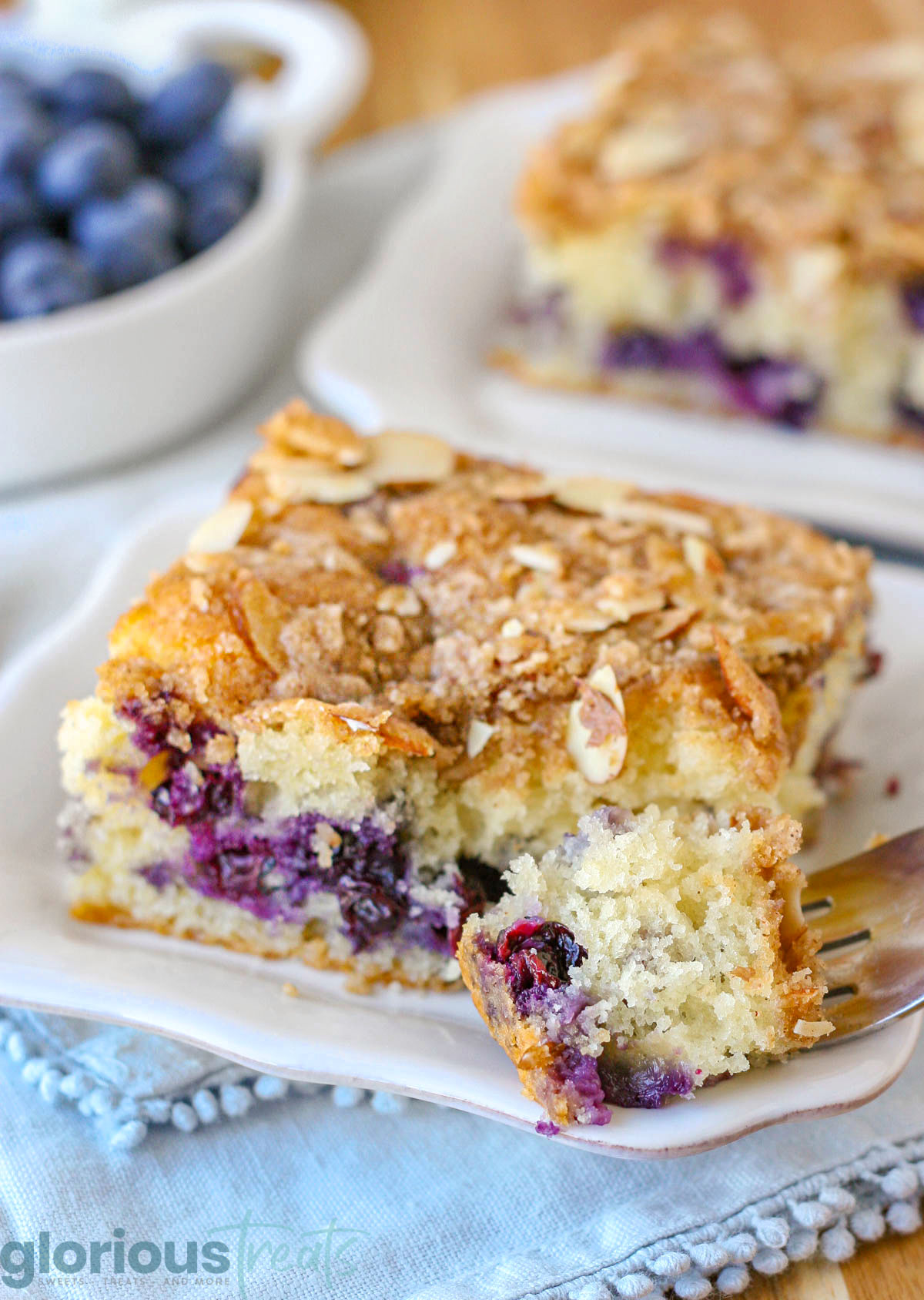 A piece of blueberry coffee cake on a white plate with a fork holding a bite sized piece.