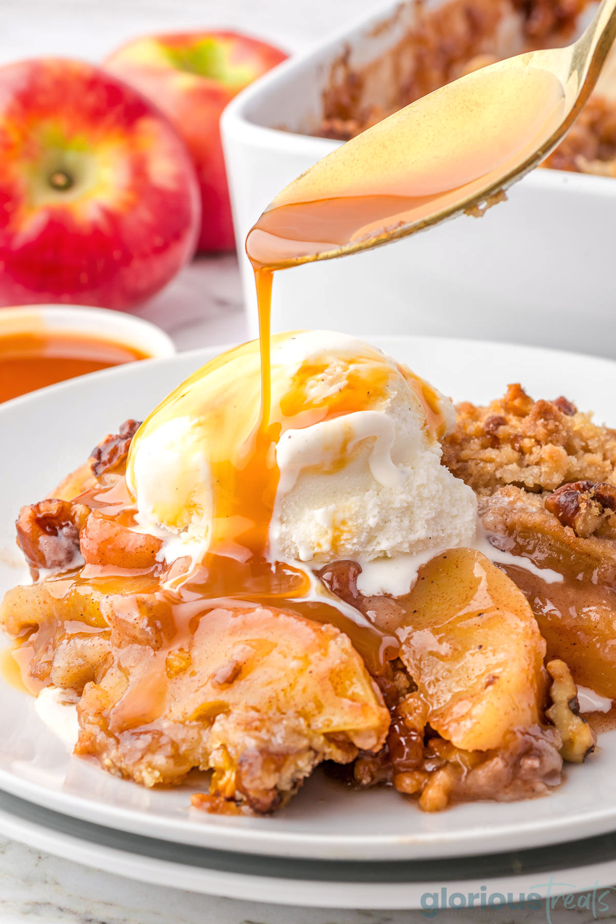 A gold spoon drizzling caramel sauce onto a serving of apple crumble on a white plate.